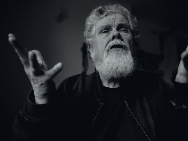 A black-and-white image of a man with a white beard and wavy white hair, gesticulating with his hands. 