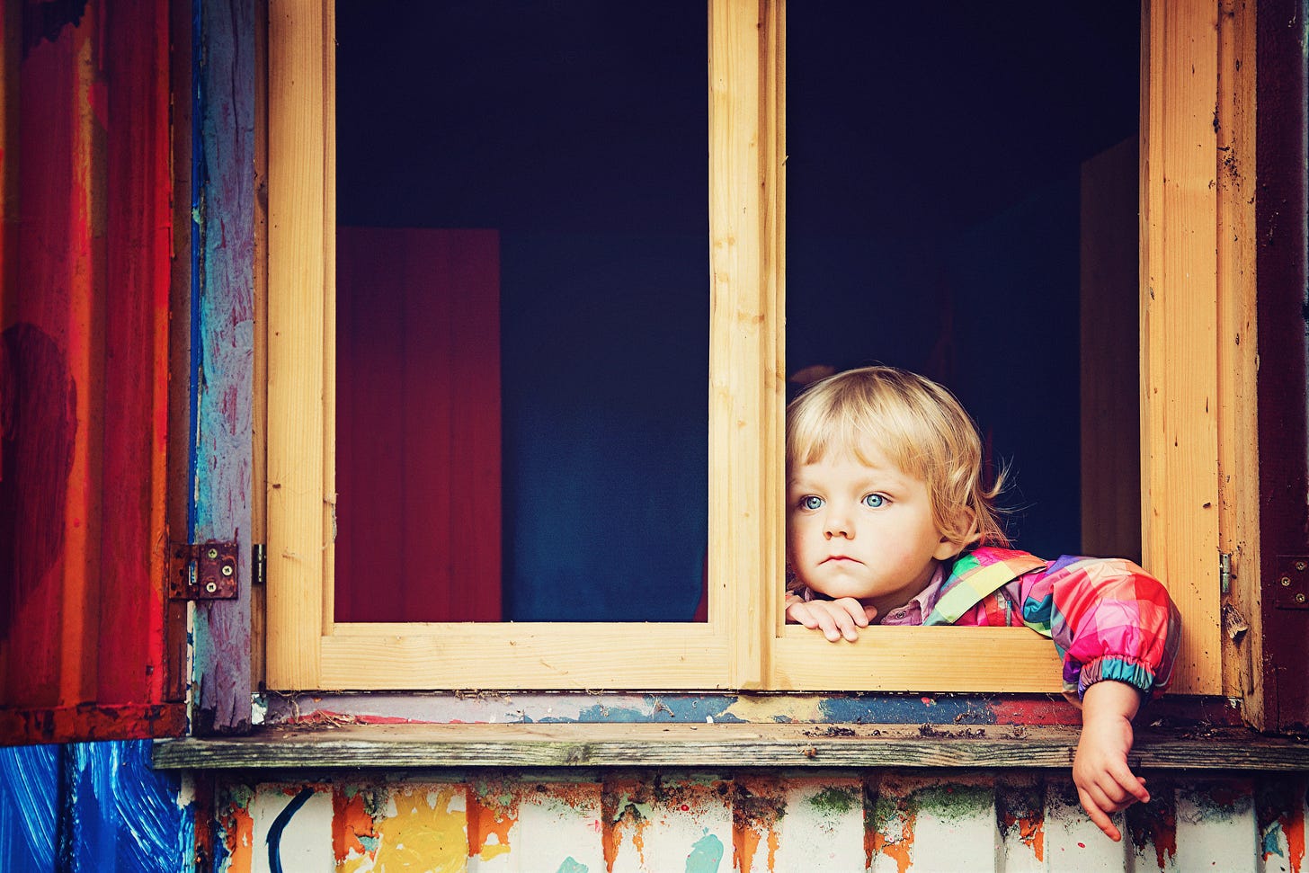 Young child sat at an open window looking longingly, sad