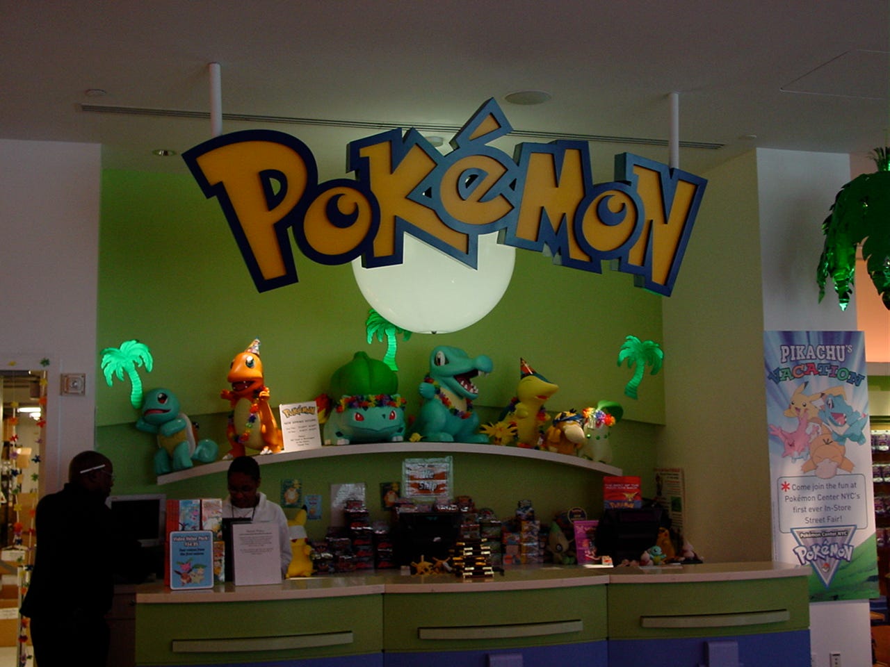 A photograph of Pokémon Center New York’s checkout area on July 22nd, 2002. The store was decorated with palm trees, flower necklaces, and party hats to celebrate the store’s “Pikachu’s Vacation” event, described as an “In-Store Street Fair”