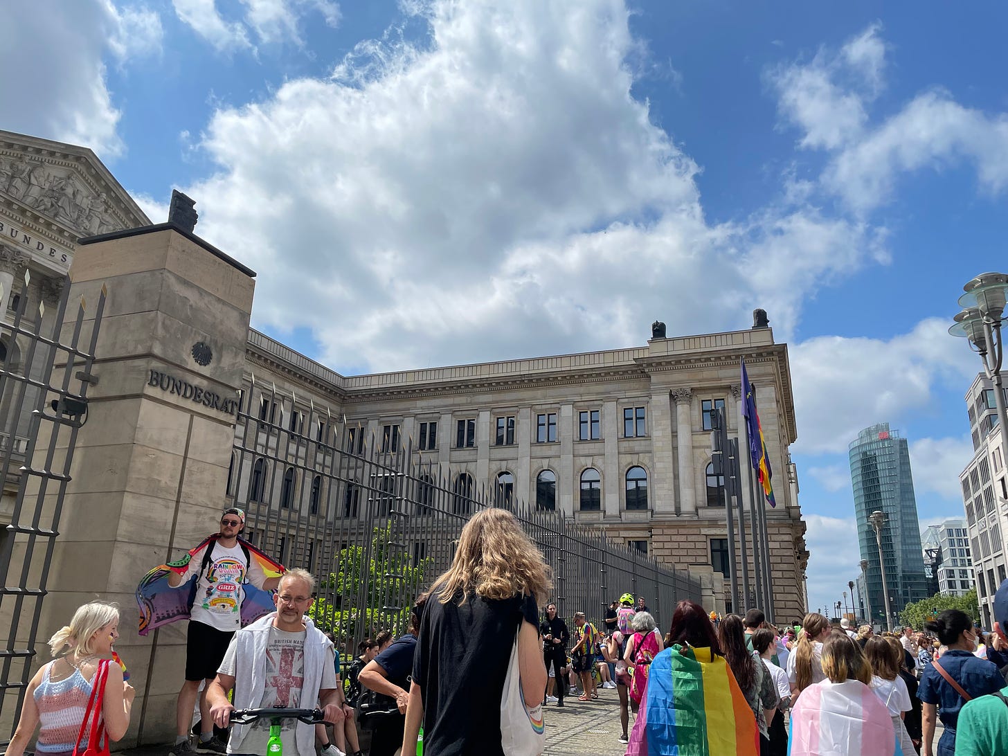 The Bundesrat building in Berlin during CSD 2023, with a crowd of people, some draped in rainbow flags, gathered outside. The Pride flag flies alongside the German flag against a backdrop of blue skies and clouds.