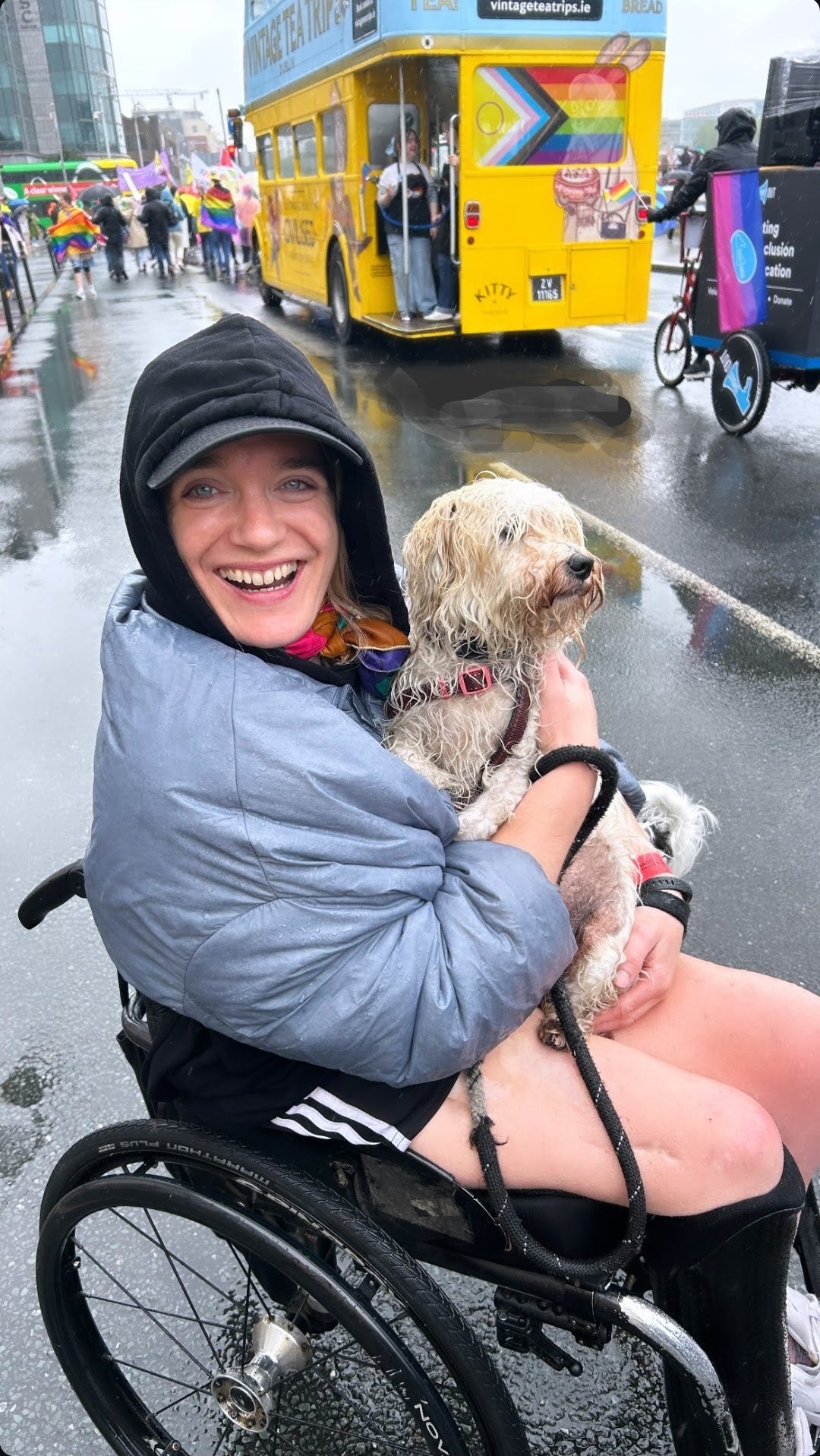 Wearing a rain coat, O'Neill's striped shorts, my hood up with a baseball cap underneath it, I am sitting in my wheelchair in the middle of the Pride Parade with my sopping wet white terrier on my lap. It is pouring rain, the roads are wet and people behind wave Pride flags in front of a double decker Vintage Tea Bus that is also kitted out in Pride flags. 