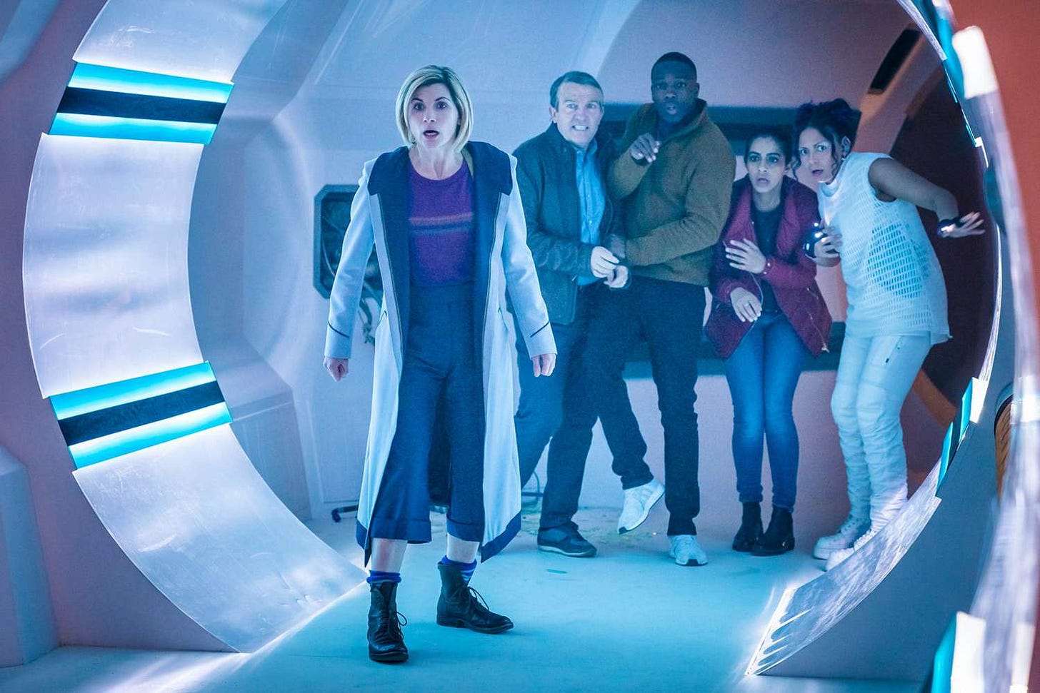 This image from "The Tsuranga Conundrum" (Doctor Who Series 11's fifth episode) shows Team TARDIS (from left to right, the Thirteenth Doctor, Graham O'Brien, Ryan Sinclair, and Yaz Khan) and Tsuranga physician Mabli (played by Lois Chimimba) standing in a brightly lit corridor of their medical ship and reacting in shock to the offscreen Pting's seeming attacks. This image from "The Tsuranga Conundrum" (Doctor Who Series 11's fifth episode) shows Team TARDIS (from left to right, the Thirteenth Doctor, Graham O'Brien, Ryan Sinclair, and Yaz Khan) and Tsuranga physician Mabli (played by Lois Chimimba) standing in a brightly lit corridor of their medical ship and reacting in shock to the offscreen Pting's seeming attacks.