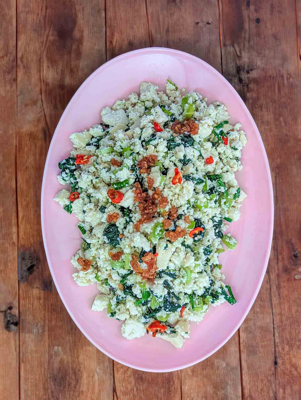 Top down photo of a pink oval plate on a rough wooden surface. The plate contains crumbled tofu mixed with chopped greens and flecks of red chillis 