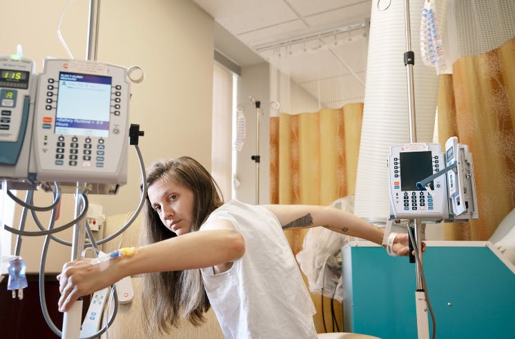 In a photograph, a white person with long brown hair is gripping the metal poles holding IV bags, seated on the edge of a hospital bed out of the shot.