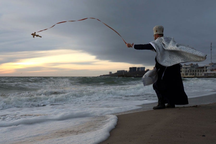 An orthodox priest tosses a small cross into the surf, holding onto a ribbon attached to the cross.