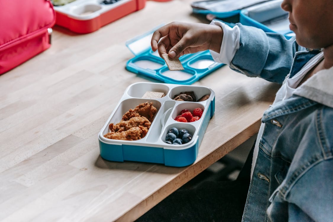 Free From above side view of crop unrecognizable ethnic schoolkid at table with lunch container full of tasty food Stock Photo