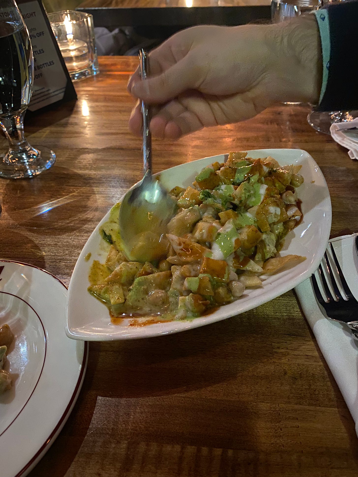 An ovoid dish of papri chaat: chickpeas and crispy roti pieces covered in yogurt sauce and a couple of different chutneys. Jeff's hand is serving some out with a metal spoon.