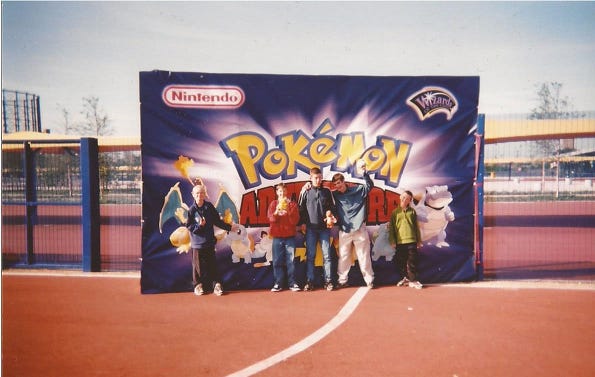 A photo of Paul and his friends standing in front of a sign with various Pokémon on it.