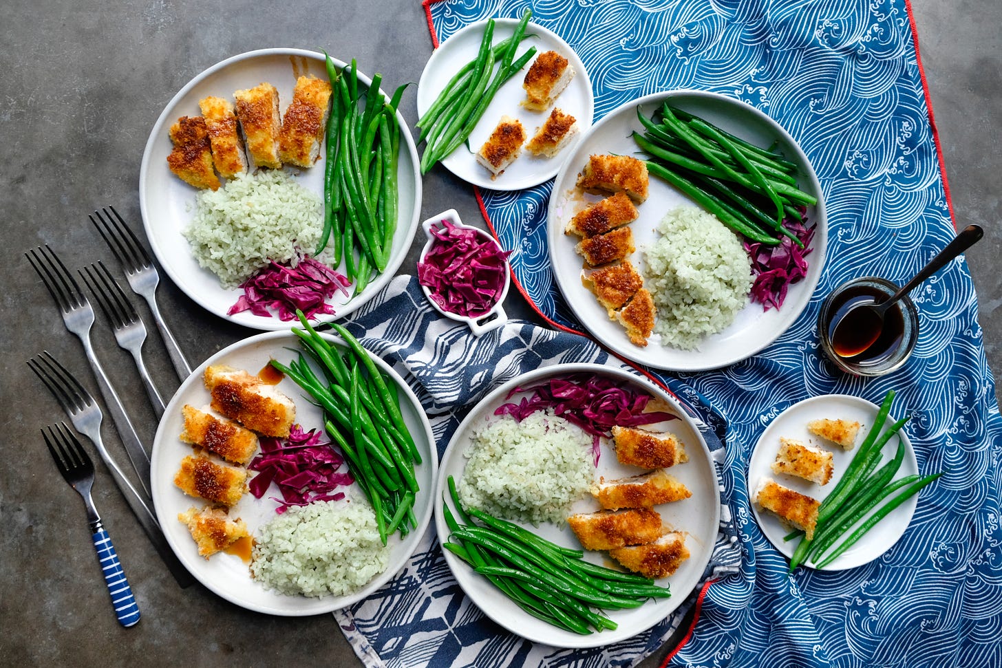 Five plates of Turkey Katsu (breaded turkey paillards) with jade rice, haricot vert, jade rice, and pickled cabbage, on a gray table with a blue tablecloth