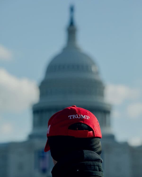 A man wearing a Trump hat stands in front of the Capitol.