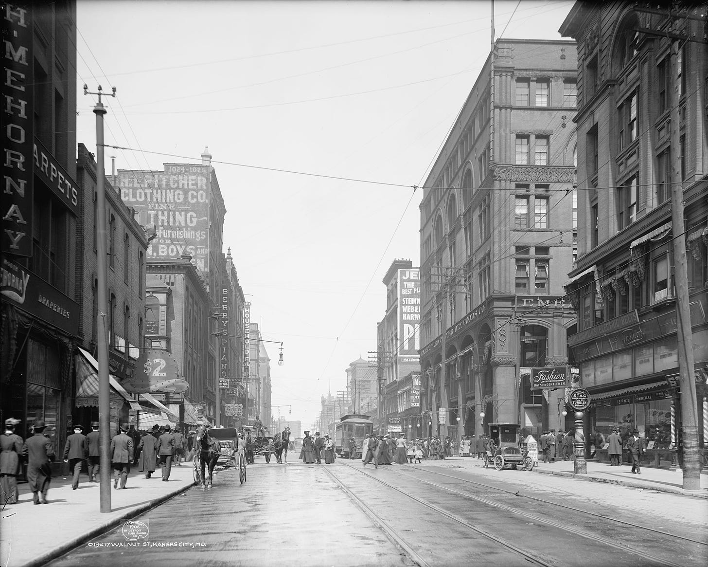 A photograph of a wide city street with large buildings, a tramway, horse-drawn vehicles and pedestrians.