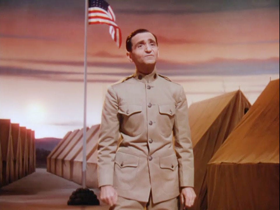 Irving Berlin in military uniform on stage during 1943 film This is the Army. He’s singing and behind him are rows of army tents against a sunset sky with the American flag fluttering at the top of a pole