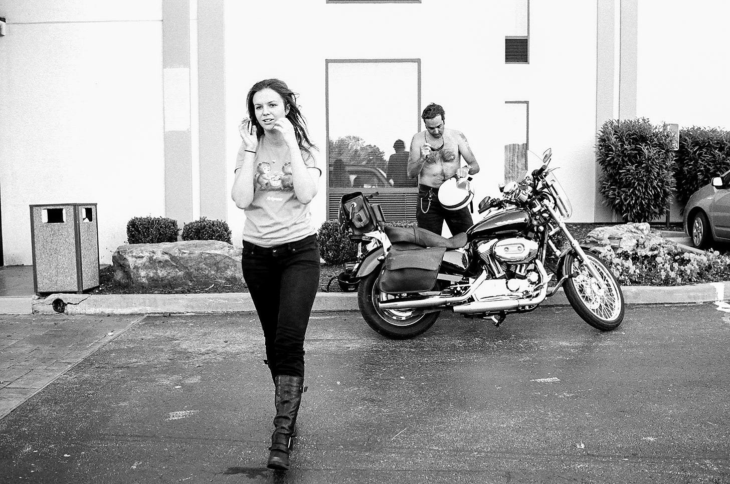 Derrick and Amber in a parking lot. Derrick is standing next to the parked motorcycle with his helmet in his hand. Amber is walking toward the camera as if she's going to pass by it. Her hands are near her face like she's about to adjust her hair from wearing her motorcycle helmet. 