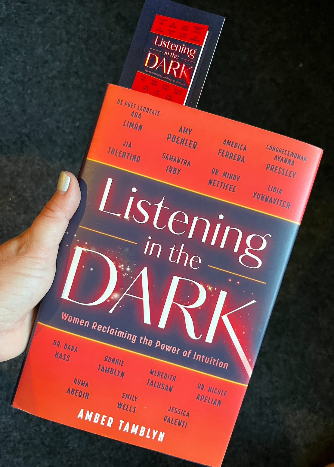 A hardcover copy of the book "Listening in the Dark: Women Reclaiming the Power of Intuition." Amber's hand holds the book in frame. A bookmark matching the cover of the book is sticking out from between the pages. 