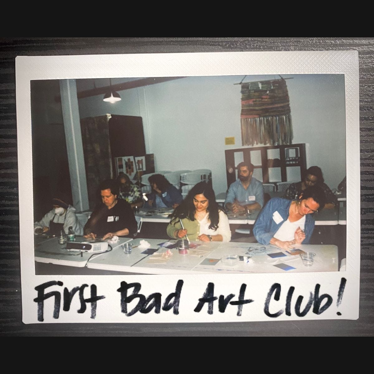 A polaroid photo of BAC with "First Bad Art Club!" written at the bottom. The photo is of 4 women working on art on the front row, and 2 men and 2 women working on art in the second row of the classroom.