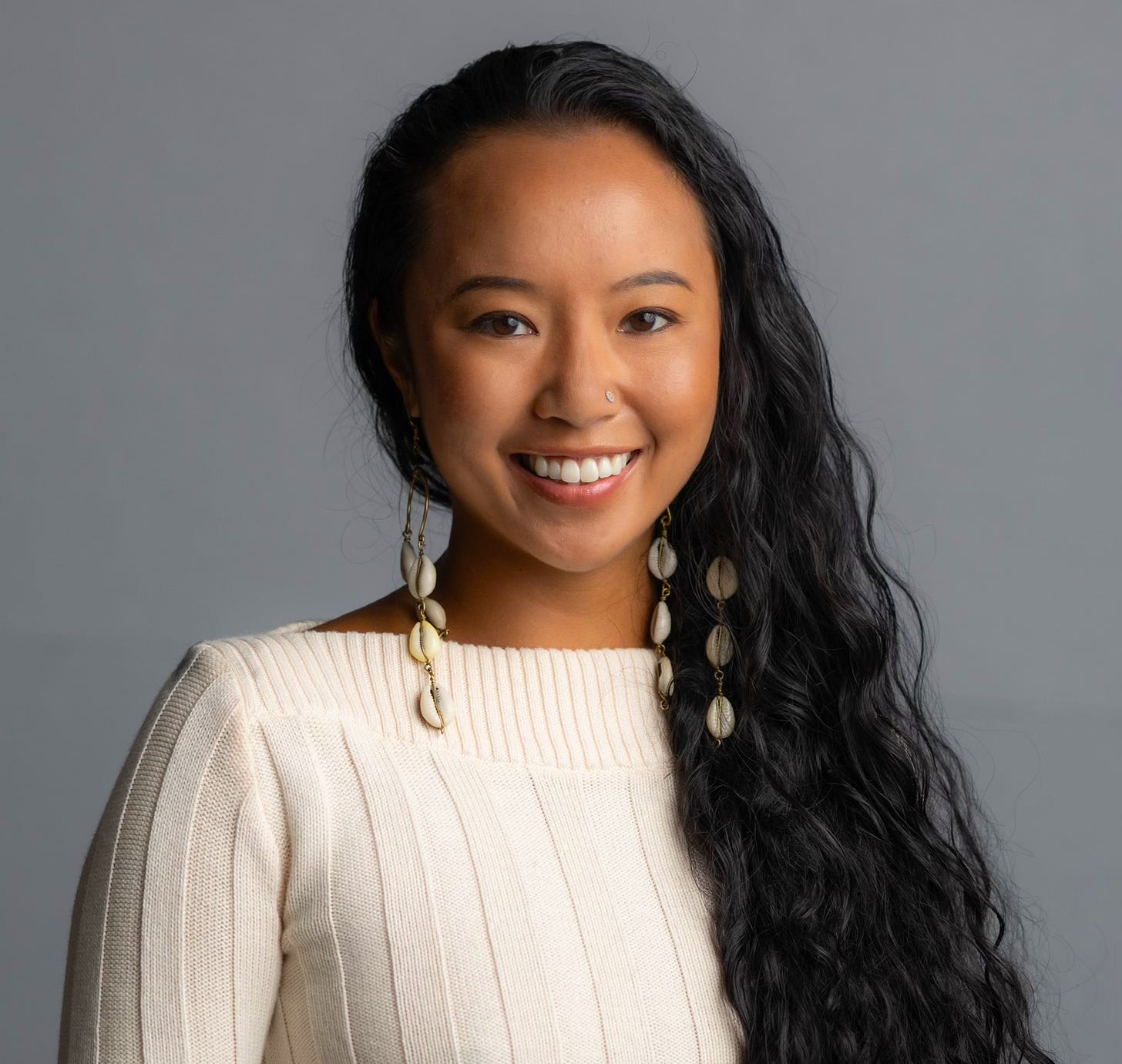 Bernie smiling, with long brown heair over one shoulder, dangling cowrie earrings and a cream colored sweater, against a grey background