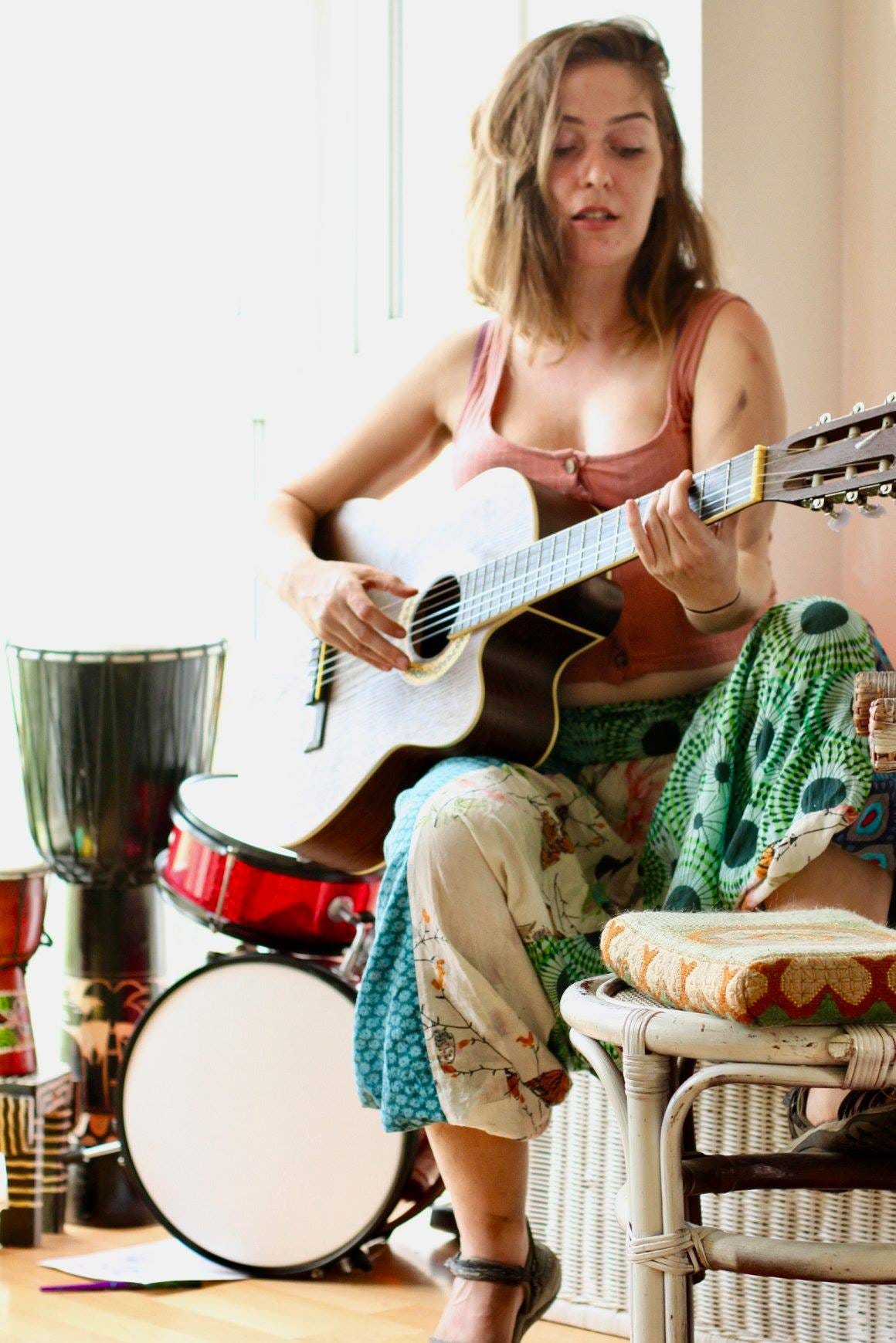 Ellese, a white woman with light brown hair, plays the guitar,  surrounded by percussion instruments. She is wearing flowy, hippy clothes.