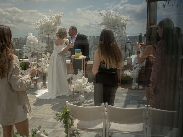 A woman in a long white, off-the-shoulder dress and a man in a black suit lean toward one another amid vases of white flowers. People are gathered around them at a distance, some taking photos.