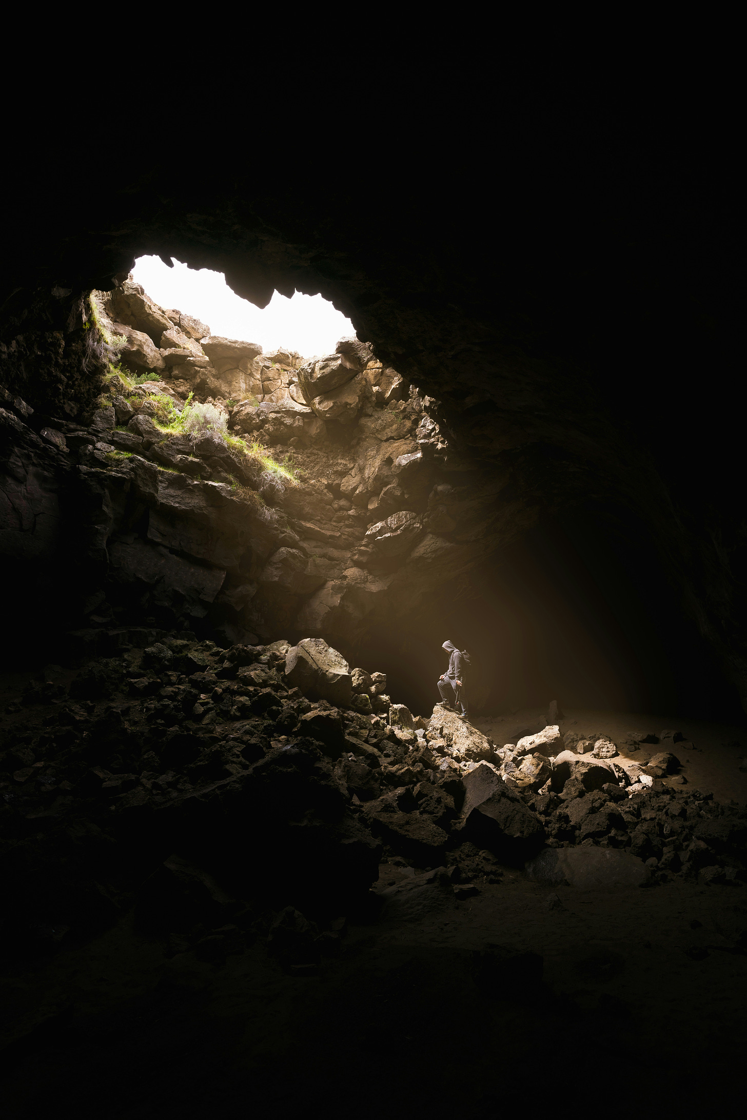 Light sreams through a large opening into an underground cave, illuminating a climber who is looking up at the outside world.