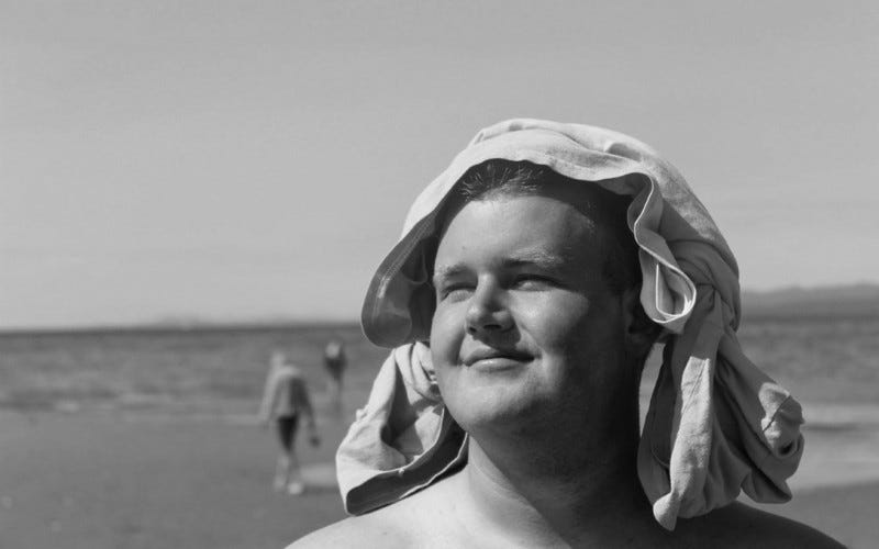 A young man on a beach, smiling, with a shirt on his head.