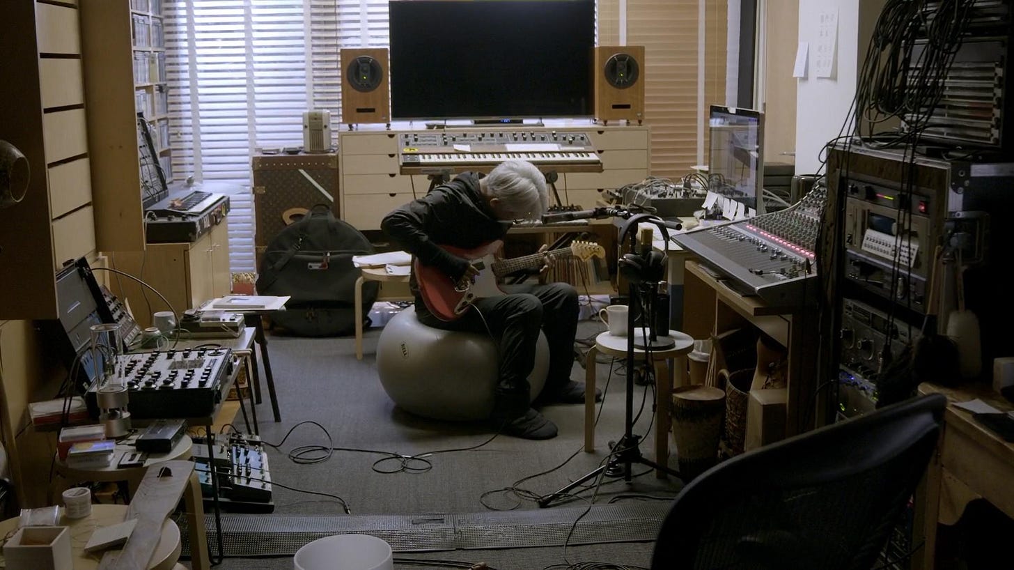 Ryuichi Sakamoto sentado numa bola cinza de pilates, tocando guitarra com a cabeça abaixada. Ele está vestido todo de preto e está cercado por fios e equipamentos de música.