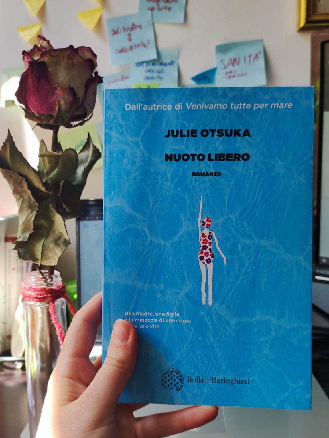 Una mano tiene un libro: Nuoto libero. La copertina è azzurra, con una nuotatatrice con un vestito a fiori al centro. Dietro il libro si intravede una rosa sulla sinistra, un computer e un muro pieno di post it