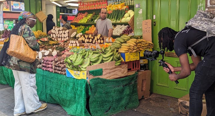 A filmmaker filming a woman browsing a fruit and veg stall in Brixton