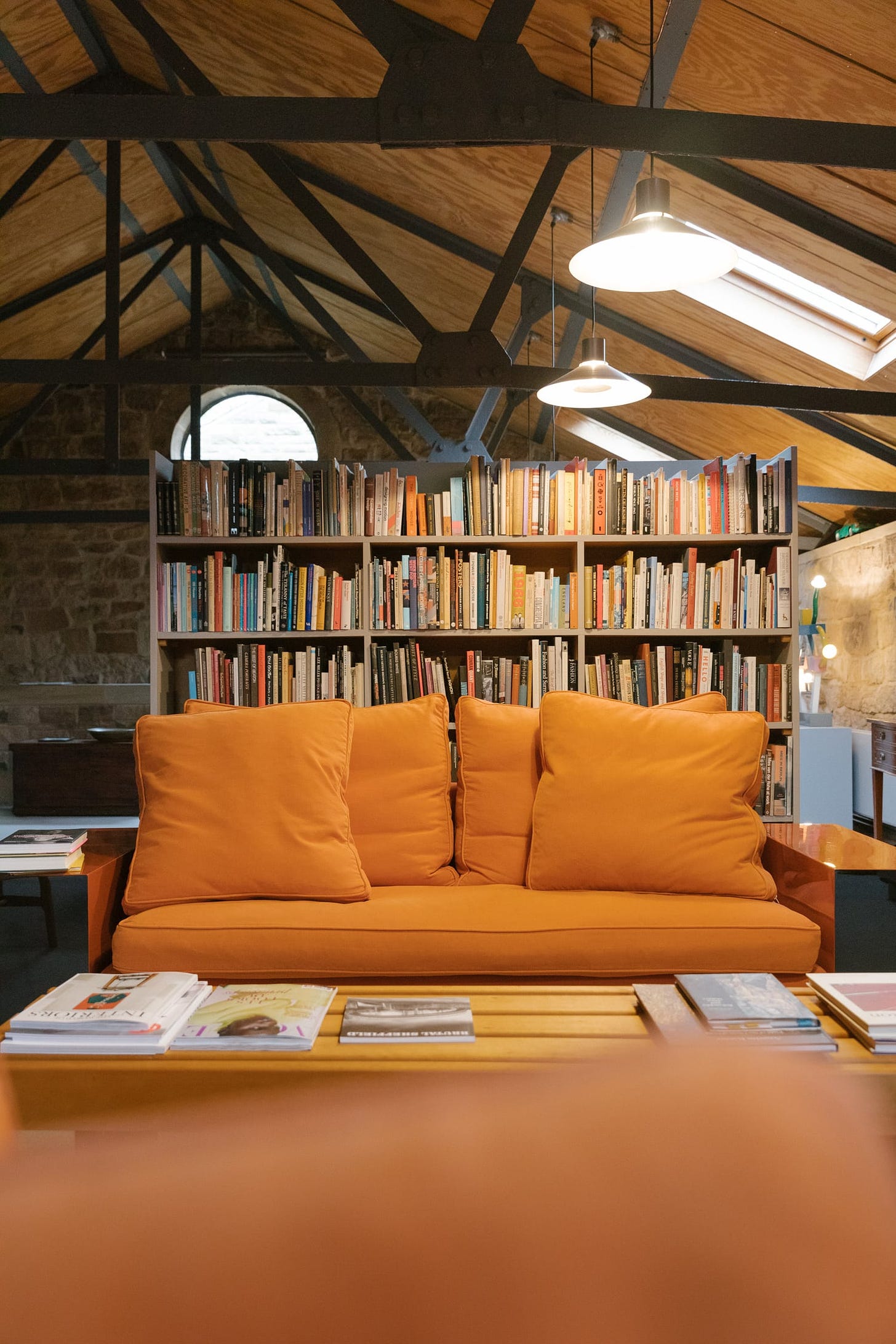A living room scene with a large 2 seater sofa. Bookshelves behind the sofa full of design and art books. Stone walls and a vaulted ceiling with steel frame.