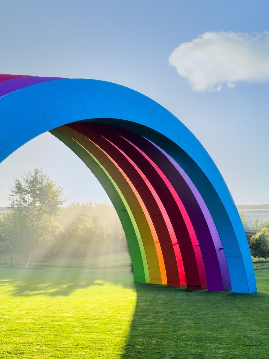 A photo of half of the rainbow at Apple Park with light beams peeking out underneath the rainbow, onto the green grass. The blue sky and trees are behind the rainbow, with one white fluffy cloud in the sky.
