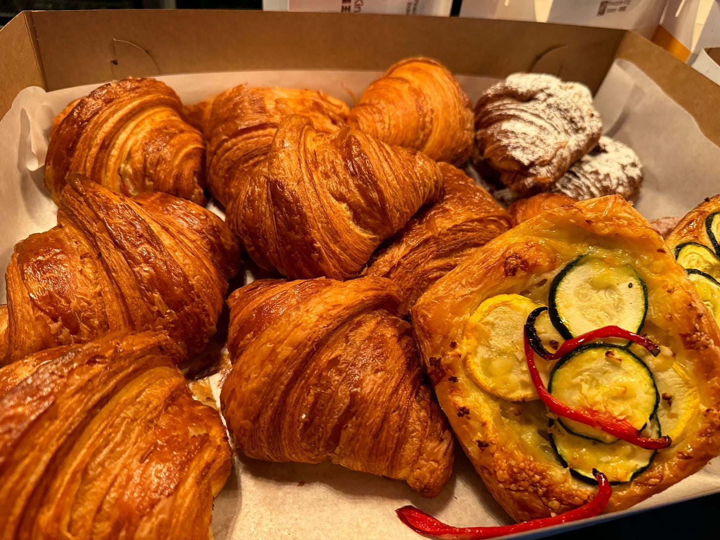 A cardboard box, lined with parchment paper, filled with croissants and other baked goods.