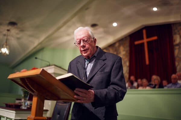 Jimmy Carter standing at a lectern reading from a book with a cross in the background.