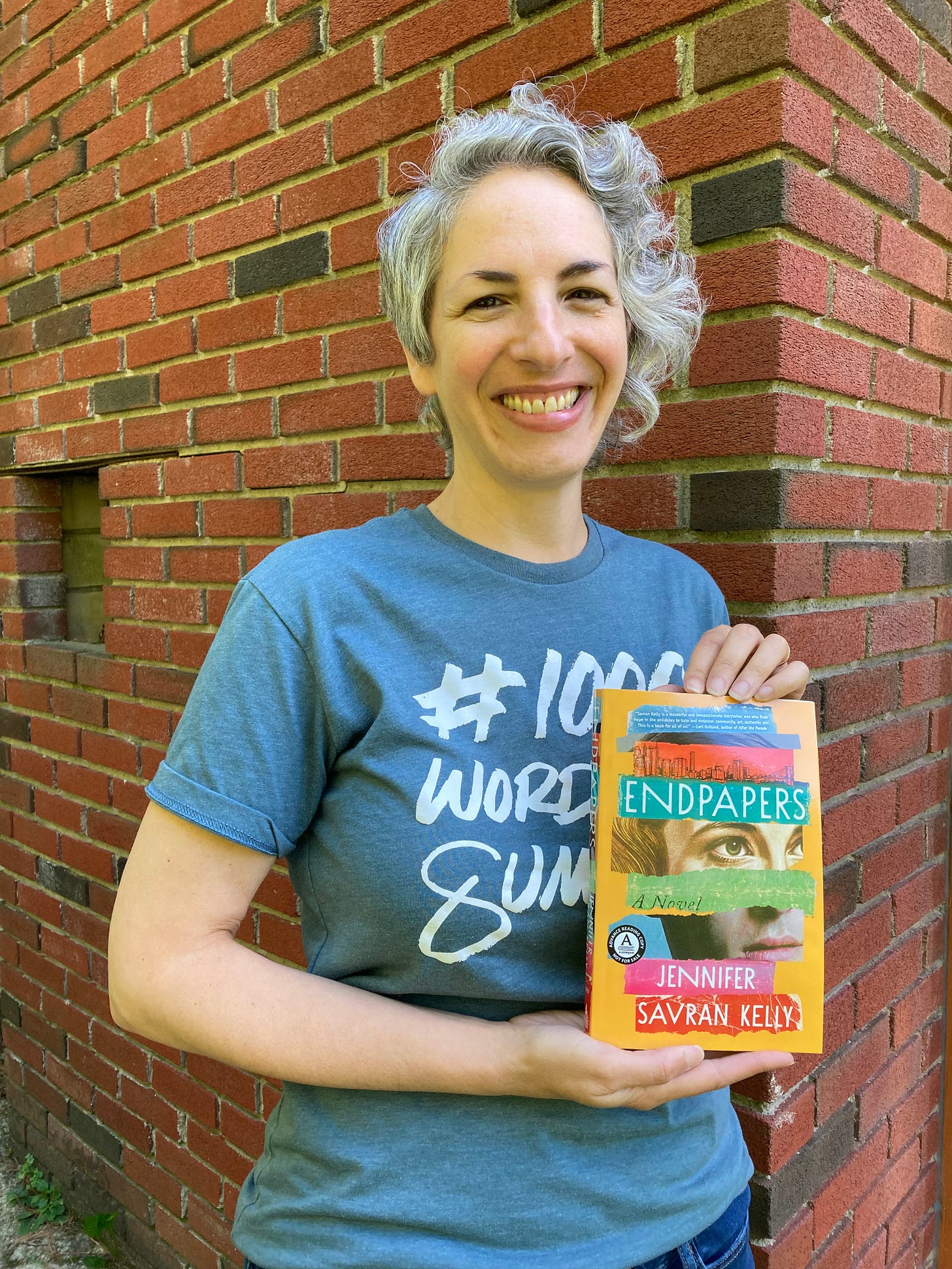 I'm standing in front of a brick wall, outdoors, in a blue T-shirt with white writing that says "#1000WordsOfSummer" and holding a copy of my novel. I'm smiling.
