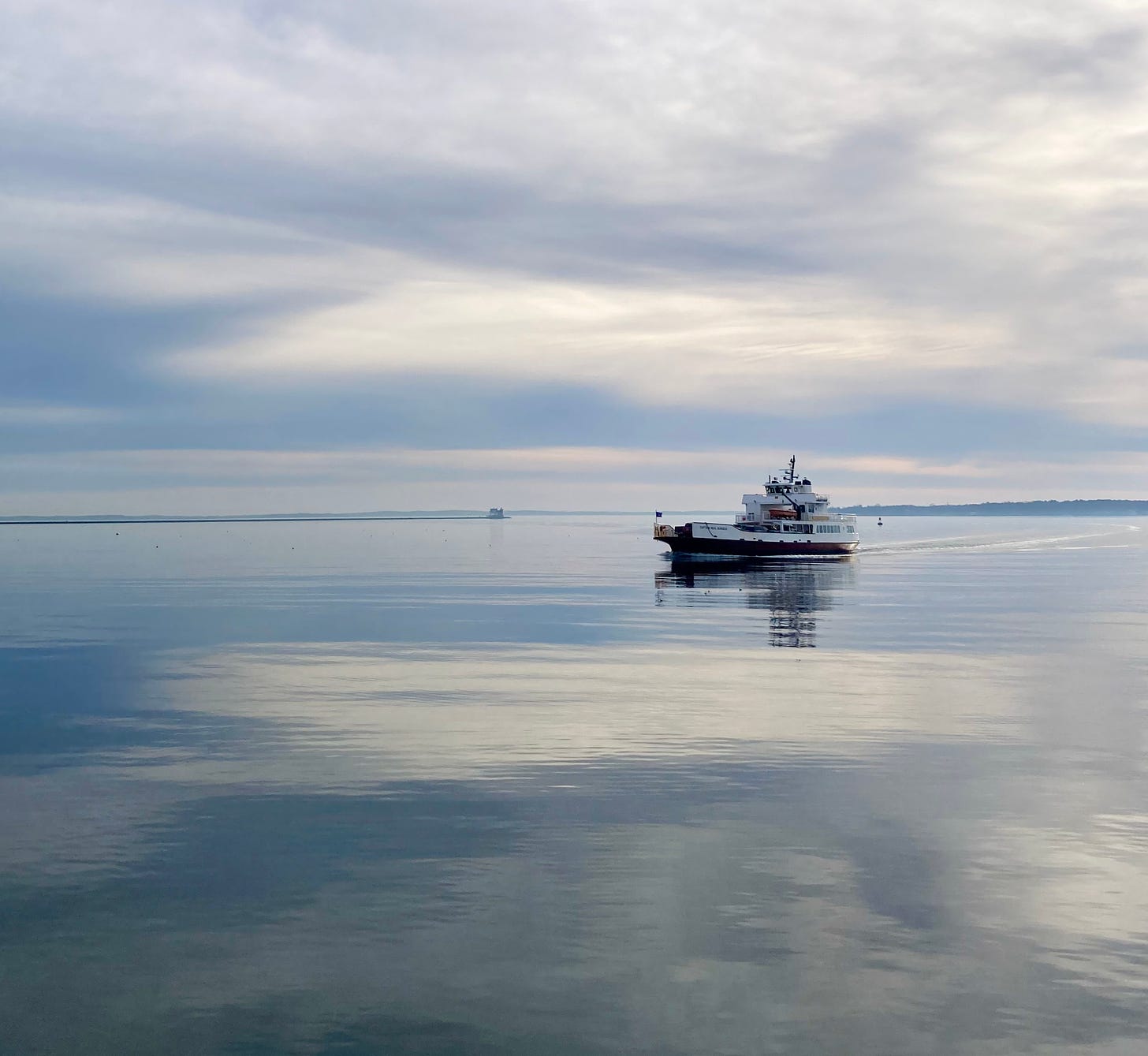 A ferry glides through calm water in a harbor. The sky is overcast and reflected in the water. A breakwater and lighthouse is visible in the background.