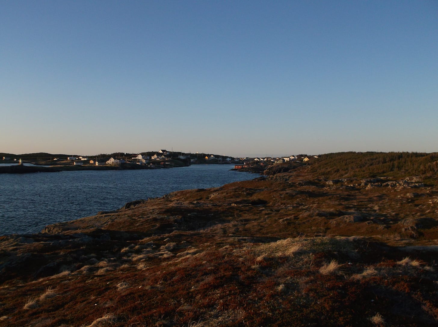 An island village at dusk. The shoreline is rocky and there are houses in the distance.