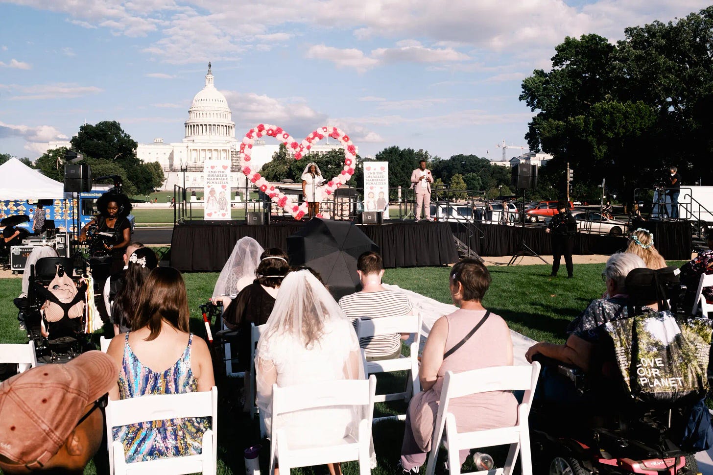 A photo of the National Mall with the Capitol building behind a stage. A wedding officiant demonstrates maximum brightness and audacity from a stage outfitted with a towering pink-and-red heart and “End the Disabled Marriage Penalties” banners. Disabled couples sit in white chairs on the lawn.
