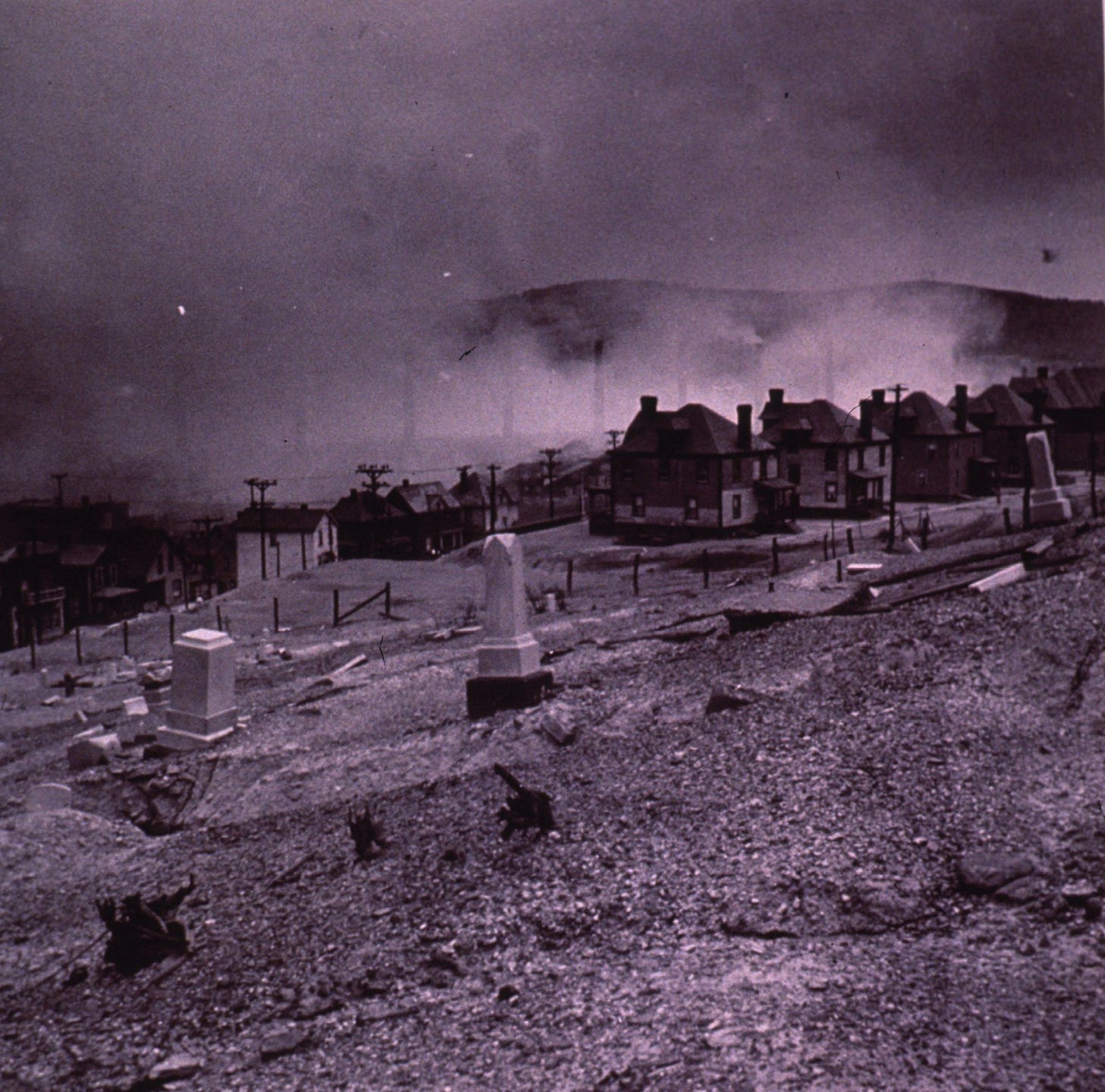 cemetery on hillside, identical houses in middle ground, smoke mostly obscuring smokestacks beyond