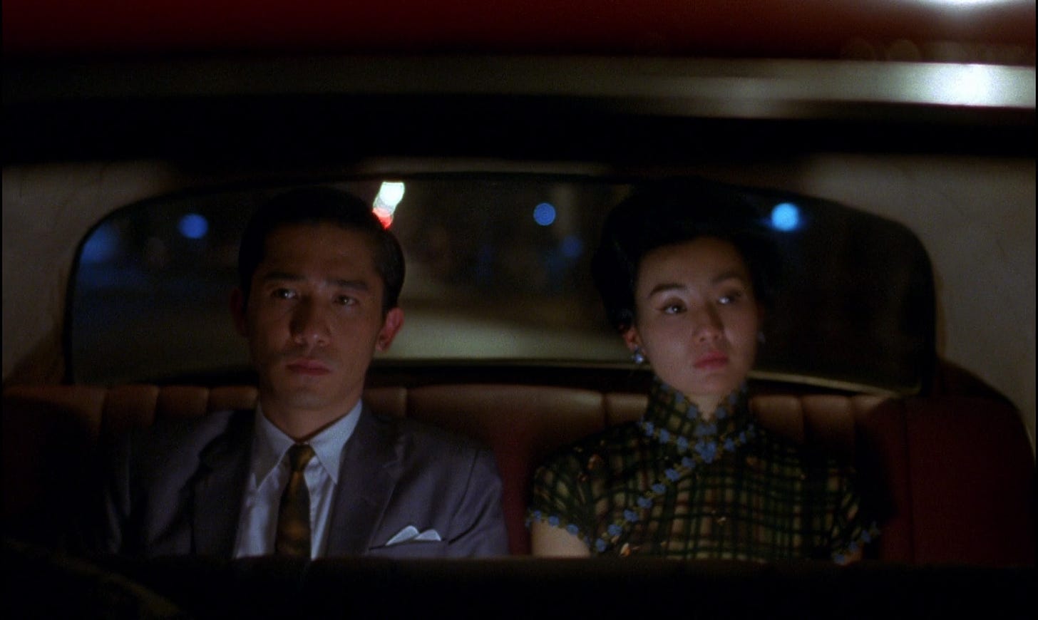 A man and a woman sit side by side in the back seat of a car, not looking at each other. It is night.