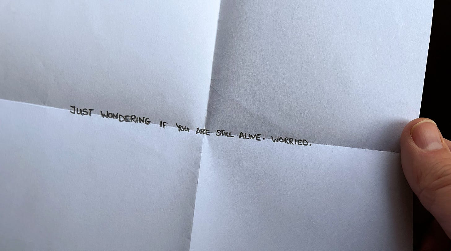 A piece of paper is being held and in the middle is a single line of text saying just wondering if you are still alive. Worried.