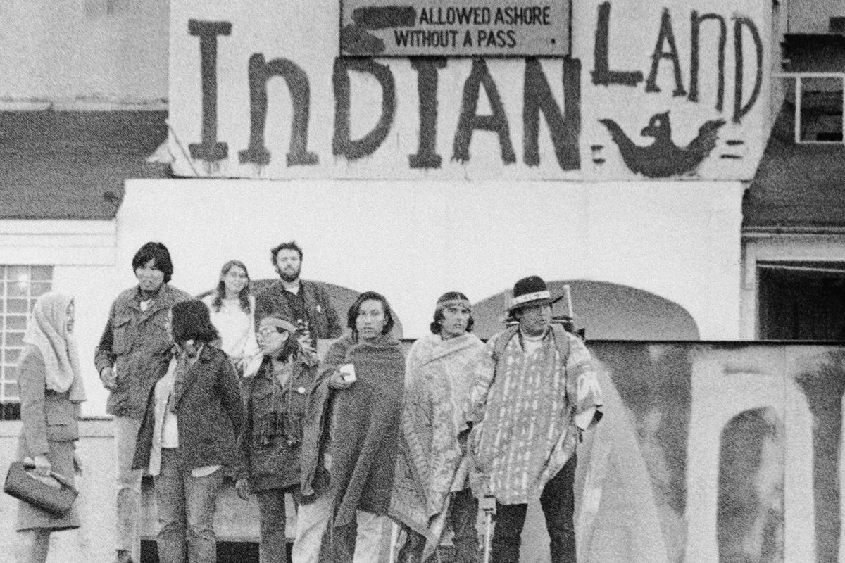 black and white photo of nine people standing in front of a wall in 1969 that reads "Indian Land" written in paint