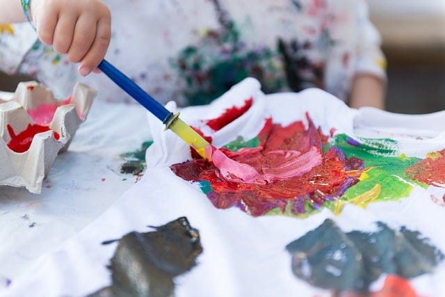 A child's hand painting a messy painting with tempera paint and a big brush. Via Pixabay, in the public domain.