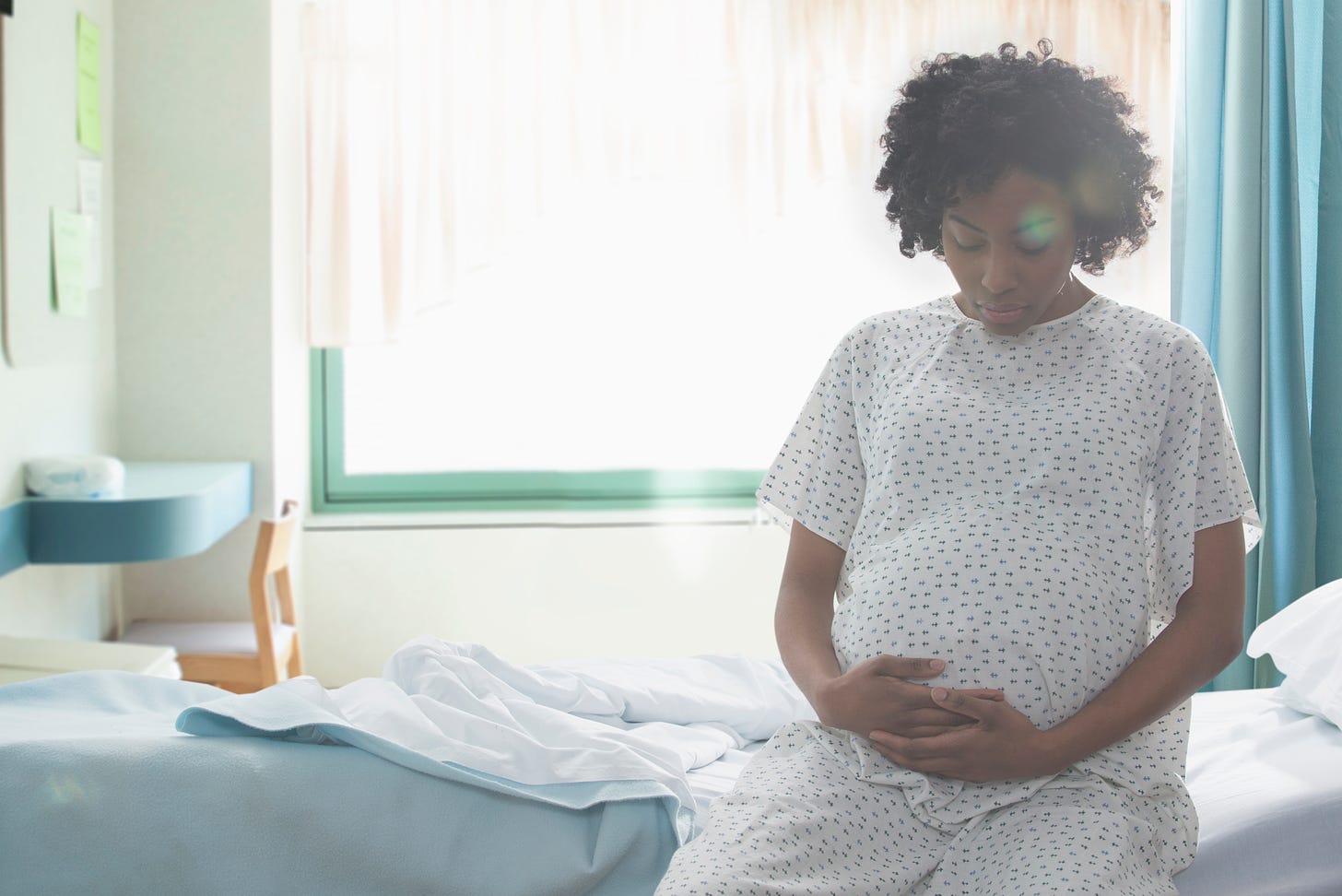 An image of a Black pregnant person in a medical gown, sitting on a hospital bed and holding their pregnant belly as they look down at it.