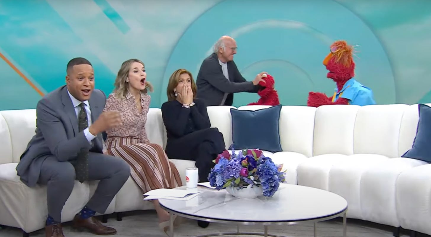 An image from Today; several interviewers sit on a wide white couch looking shocked, as behind them, Larry David grabs Elmo by the face