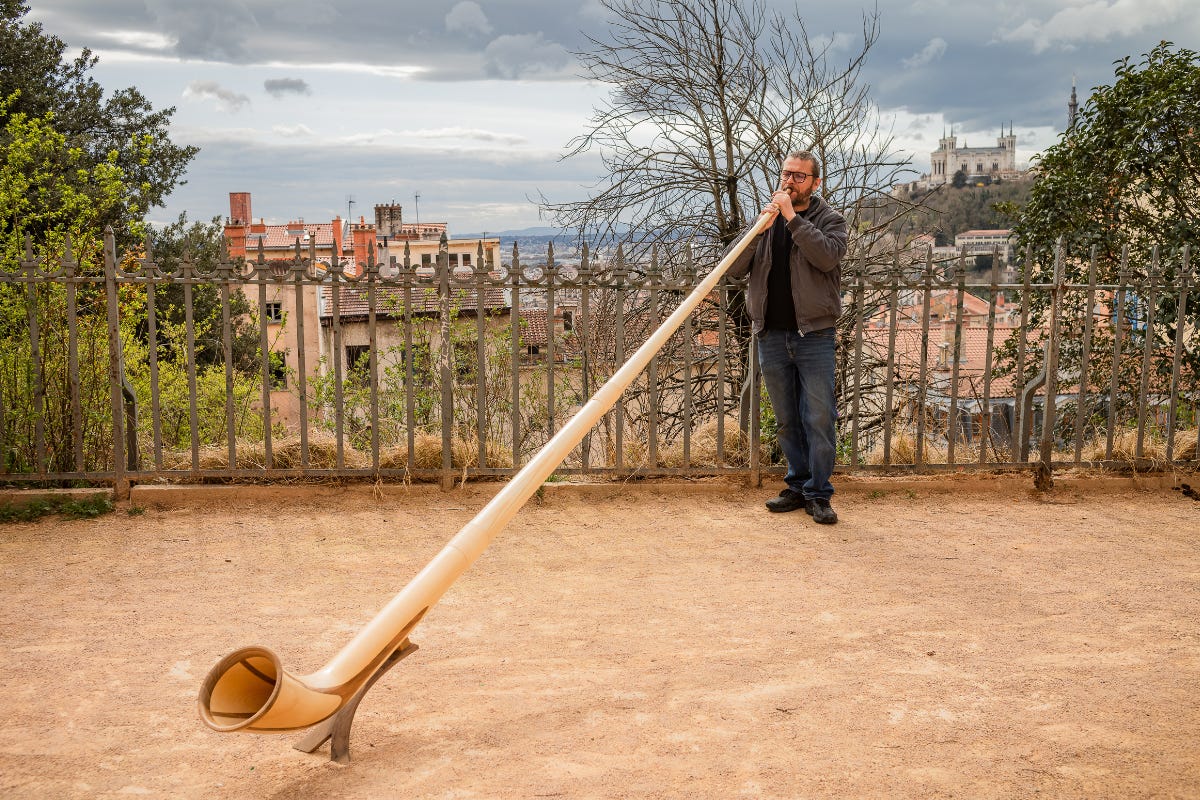 Sandro Faïta stands in front of an iron fence and plays an alphorn, with a cityscape and hillside visible in the background.
