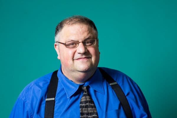 Steve Silberman poses for a portrait. He is wearing a cobalt blue shirt and black suspenders and standing in front of a teal backdrop.