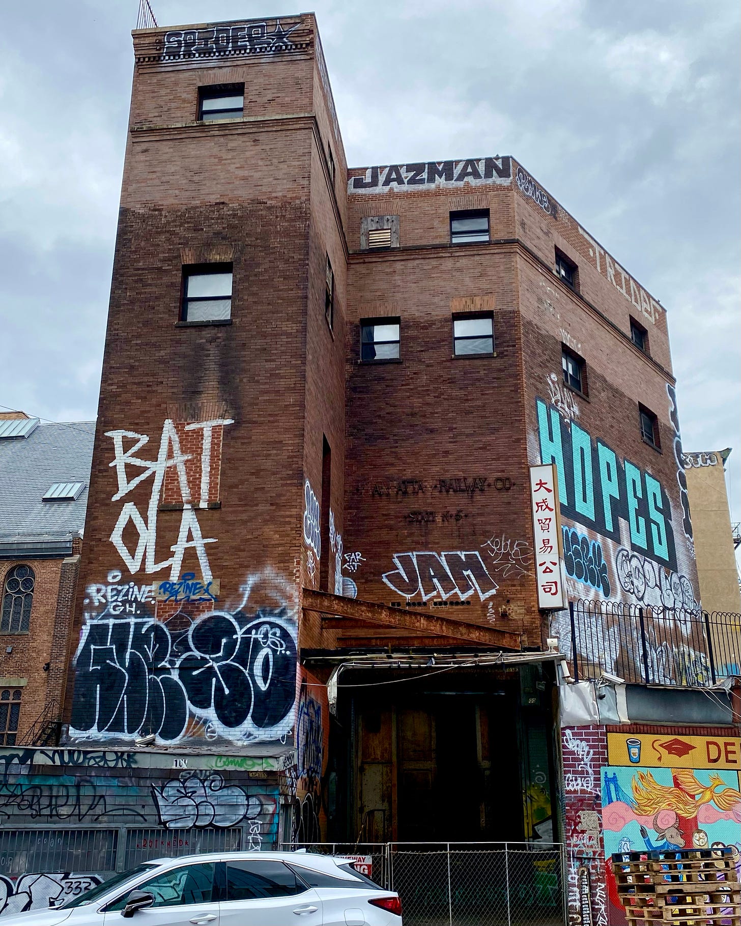 A brick building covered in graffiti. There is a sign in Chinese on one corner. Letters are missing, but you can still make out that this was marked Manhattan Railway Co.