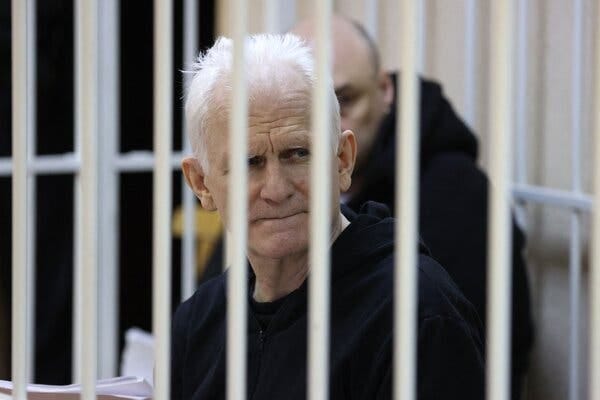 Ales Bialiatski, dressed in black, sits behind the white bars of a courtoom defenants’ cage.