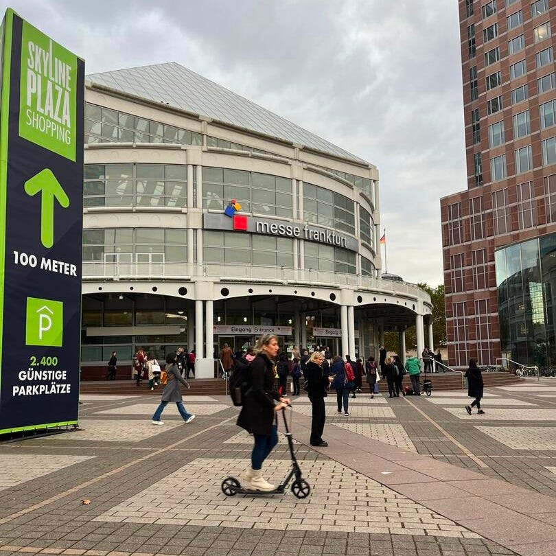 The entrance to the Frankfurt Book Fair which is a big building made of glass in front of a paved road. There are people walking around and a person on a scooter.