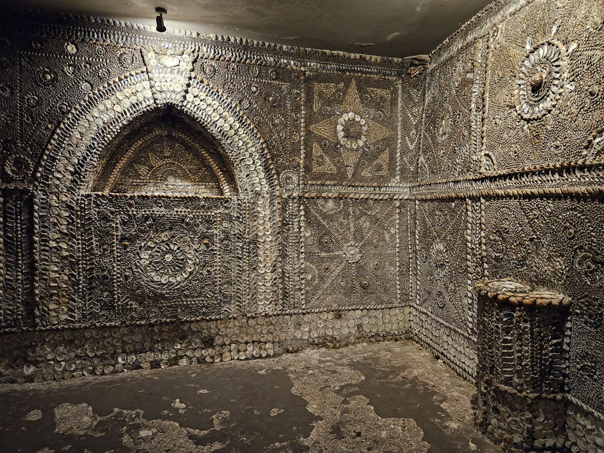 A corner of a room, with an altar on the left wall and a pedestal against the right wall, all entirely covered in shells