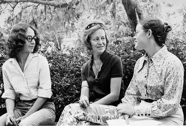 Three women sit outside together under a tree in a black-and-white photograph.