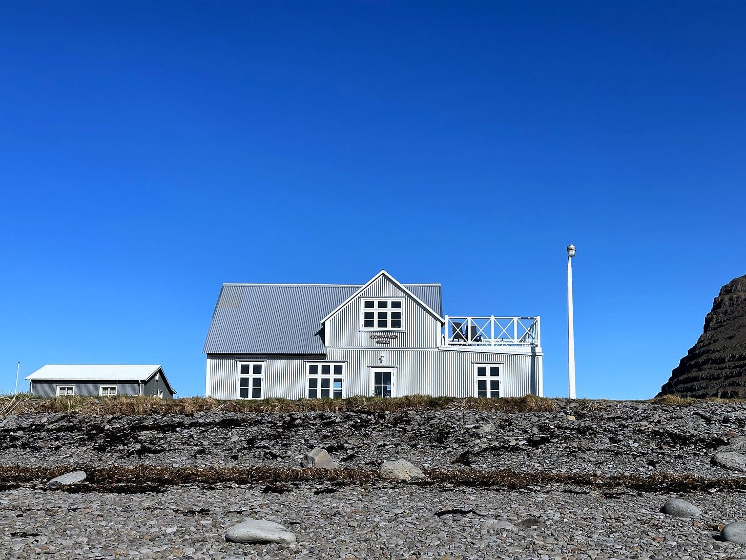 Our rental home for the night as seen from the beach.  A two storey home with silver metal siding and white windows.  The beach is covered in flat gray rocks and the sky is a cloudless bright blue.